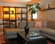 Living room area with wrap around beige couch and pillows, dark coffee table with vase and books, plant decor, open bookcase, and shelf on wall.